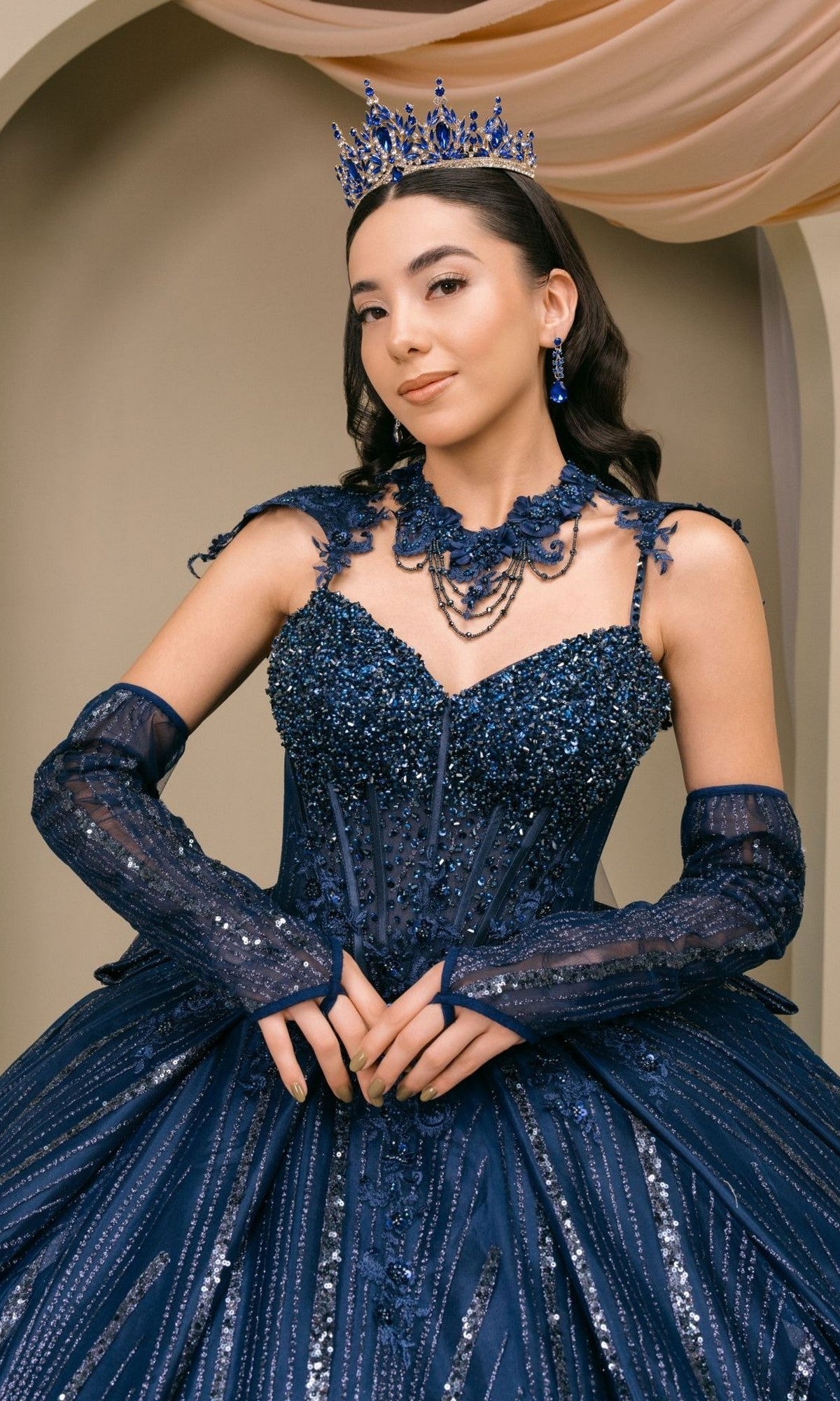 A woman wearing the elegant Quinceanera Dress 8510JQ by Cinderella Coutureโa dark blue sequin-embellished ball gownโpaired with matching gloves, a blue jeweled crown, and earrings, stands indoors against a neutral backdrop.